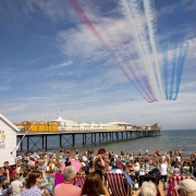 picture showing a seafront and beach filled with people with the Red Arrows flying over head