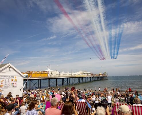picture showing a seafront and beach filled with people with the Red Arrows flying over head