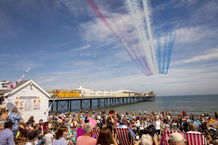 picture showing a seafront and beach filled with people with the Red Arrows flying over head